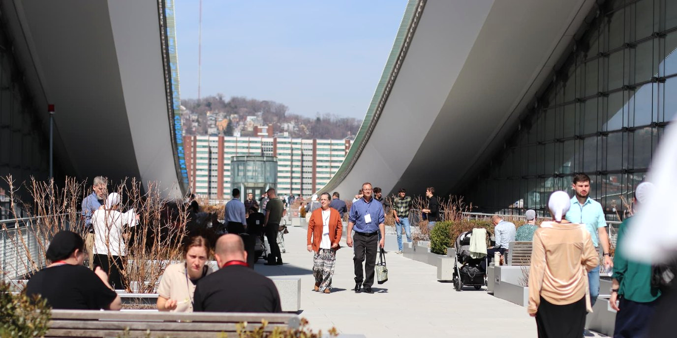 Attendees walking along a sunlit terrace between the sweeping curved canopies of the Pittsburgh convention center